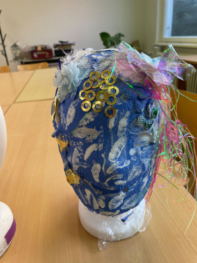 Eine Maske ist auf einem Frisierkopg angebracht. Die Maske ist blau mit weißen Tupfern. Auf der Stirn sind verschiedene Dekoelemente wie goldene Ringe, Plastikblumen angebracht.