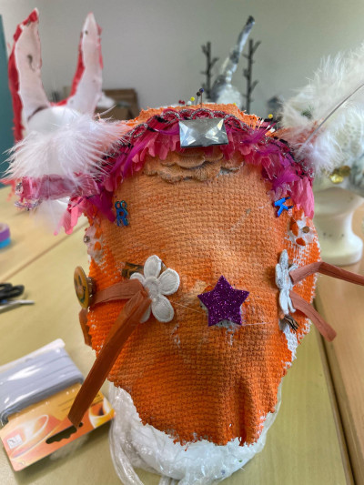 Auf einem Frisierkopf ist eine noch nicht fertige Maske angebracht. Die Maske ist orange und mit einigem Zierrat beklebt. Auf der Stirn ist eine Art Haarreif mit pinken Federn.