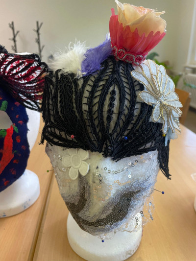 Auf einem Frisierkopf ist eine Maske angebracht. Die Maske besteht auf durchsichtigem Tüll mit Glitzer. DIe Augenpartie ist bedeckt mit einer Art venedischer Maske aus schwarzer Spitze. Auf der Stirn befinden sich Blumen.