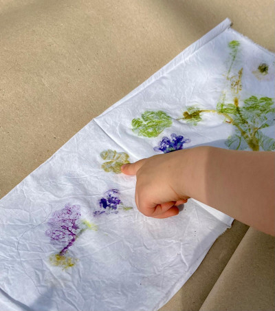 Eine Kinderhand zeigt auf einen weißen Stoff, auf dem lila und grüne Blüten gedruckt wurden.