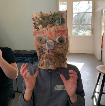 Kind trägt eine selbstgebastelte Maske aus Papiertüte mit aufgemaltem Gesicht und natürlichen Materialien wie Ästen und Blättern