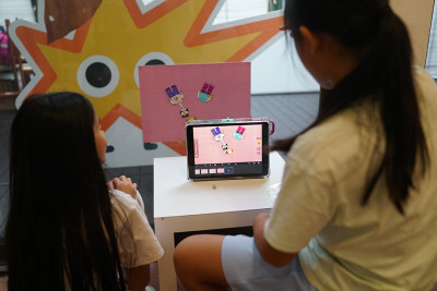 Zwei Schülerinnen betrachten ein Tablet, auf dem eine Stop-Motion-Animation mit bunten Figuren zu sehen ist