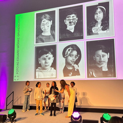 Preisverleihung des Deutschen Jugendfotopreises mit einer Gruppe von Personen auf der Bühne vor einer Projektion historischer Portraitfotos
