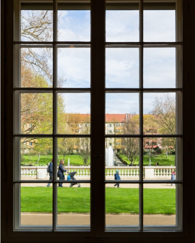 Blick durch ein großes Sprossenfenster auf den Körnerpark mit grüner Rasenfläche, Springbrunnen und Bäumen.