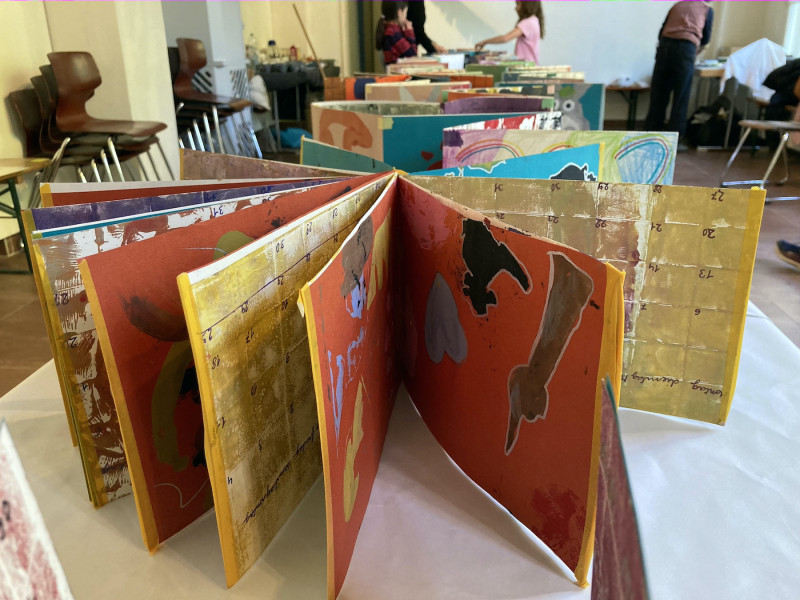 text: >
  Ein geöffnetes, farbenfrohes Kunstbuch
  mit abstrakten Zeichnungen und Collagen
  liegt auf einem Tisch; im Hintergrund
  sitzen Menschen in einem Workshop.
decorative: false
source: manual