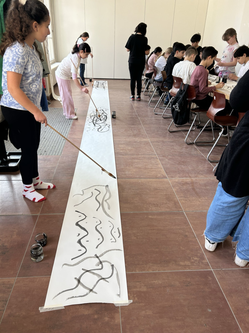 text: 'Vier Schüler arbeiten in einem Klassenraum an großen Papierbahnen: Links malen einige mit langen Stöcken schwarze Linien auf den Boden, rechts sitzen andere an Tischen und gestalten eigene Kunstwerke.'
decorative: false
source: manual