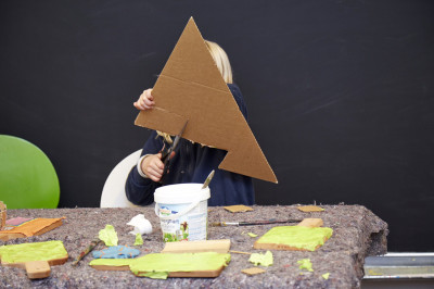Kind hält große dreieckige Pappe vor dem Gesicht während es an einem Tisch mit Bastelmaterialien arbeitet