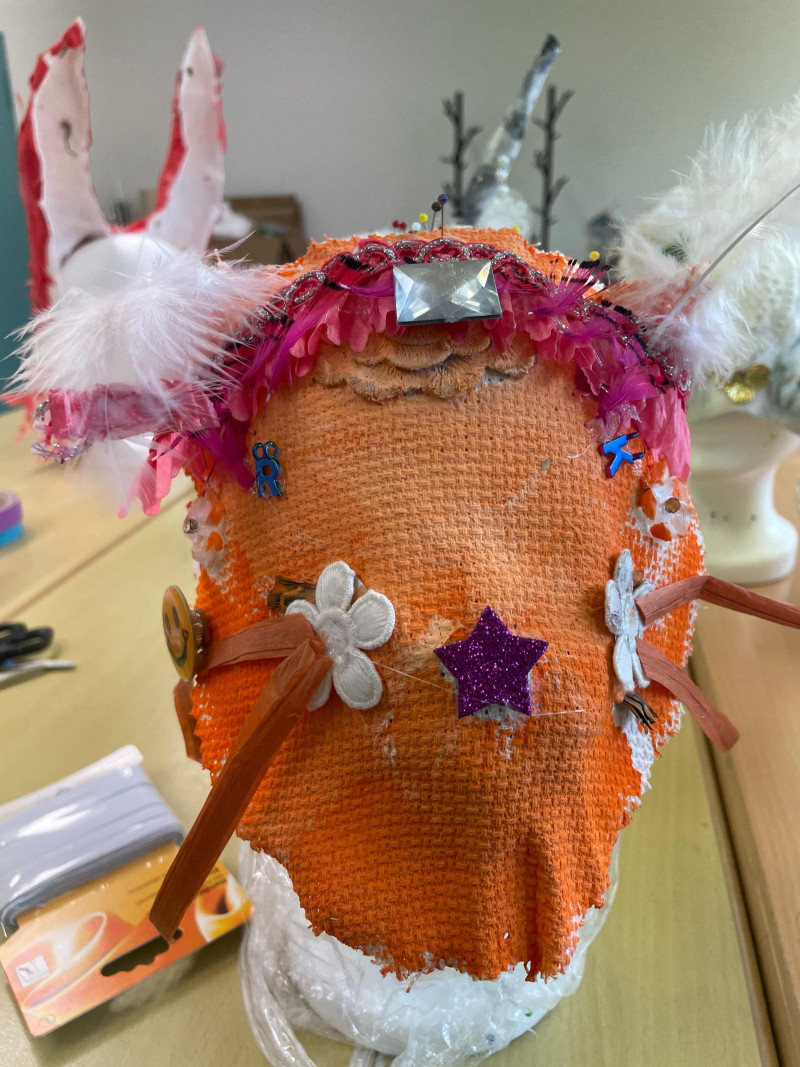 text: >
  Auf einem Frisierkopf ist eine noch
  nicht fertige Maske angebracht. Die
  Maske ist orange und mit einigem Zierrat
  beklebt. Auf der Stirn ist eine Art
  Haarreif mit pinken Federn.
decorative: false
source: manual