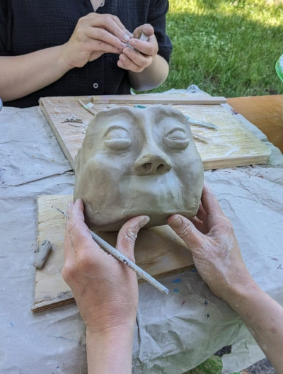 Zwei Hände formen eine Tonmaske mit ausdrucksvollem Gesicht während eines Keramik-Workshops im Freien