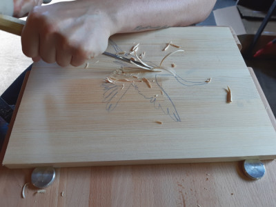 Eine Hand hält ein Schnitzeisen und schnitzt in ein Brett. Auf diesem ist ein Vogel mit Bleistift als Vorlage gezeichnet.