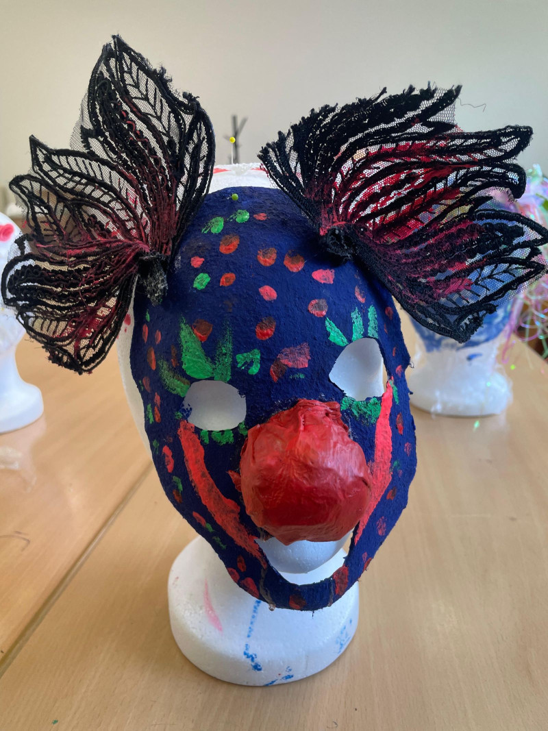 text: >
  Eine dunkelblaue Maske mit bunten
  Tupfern hängt auf einem Frisierkopf.
  Die Maske hat eine rote CLownsnase und
  an beiden Seite Federn als Deko
  angeklebt.
decorative: false
source: manual