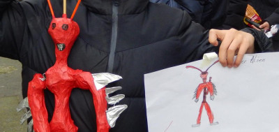 Kind bastelt rote Alien-Skulptur und zeichnet dazu passende Figur auf Papier