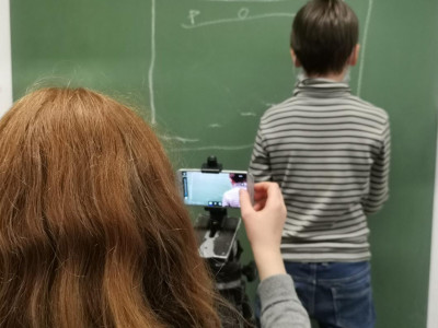 Eine Person filmt mit dem Smartphone ein Kind vor einer grünen Tafel in einem Klassenzimmer