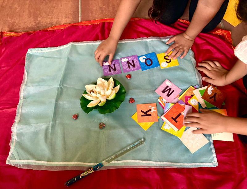 text: 'Zwei Kinderhände legen bunte Buchstabenkarten auf ein blaues Tuch, das über rotem Stoff liegt. Die quadratischen Karten in Lila, Gelb, Orange und Pink zeigen große Buchstaben sowie kleine Tierillustrationen; sichtbar ist die Anordnung **SONN**. Daneben liegt ein Stapel weiterer Karten. In der Mitte steht eine dekorative Lotusblüte mit grünen Blättern, umgeben von kleinen roten Schmuckelementen und einem glitzernden Stab.'
decorative: false
source: manual