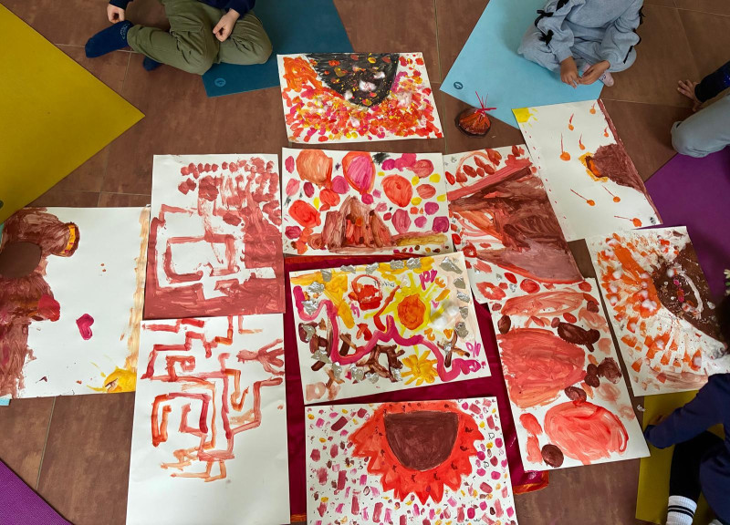 text: >
  Mehrere große Papierbögen liegen auf
  dem Boden, dicht bedeckt mit kräftigen
  abstrakten Farbflächen in Rot, Orange,
  Braun, Gelb und etwas Pink. Die
  Malspuren wirken wie breite
  Pinselstriche und Fingerabdrücke.
  Rundherum sitzen einige Personen auf
  bunten Matten und betrachten oder
  begleiten das kreative Geschehen.
decorative: false
source: manual