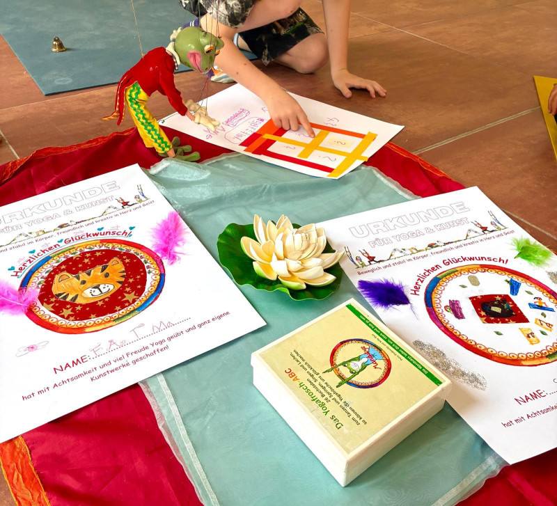 text: 'Auf dem Boden liegen zwei Urkunden für Yoga & Kunst, bunt verziert mit Federn und Mandala‑Elementen. Daneben sitzt ein Kind und zeigt auf ein Zahlenblatt, während eine grüne Handpuppe und eine Lotusblüte Teil der kreativen Szene sind.'
decorative: false
source: manual
