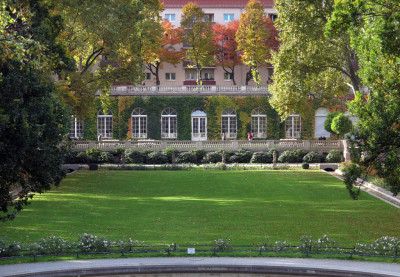 Der Garten wirkt ruhig und gepflegt: Eine große grüne Rasenfläche, umrahmt von Bäumen, führt zu einem eleganten Gebäude mit hohen, bogenförmigen Fenstern und etwas Efeu an der Fassade.